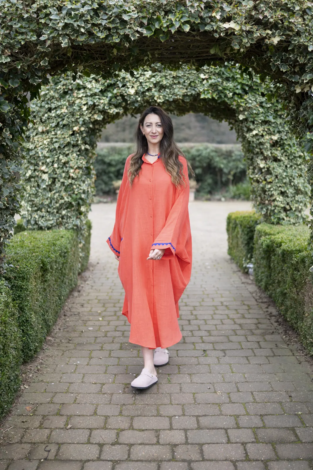 Lauren Vaknine in a coral kaftan, walking under an ivy archway